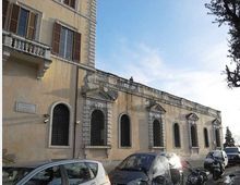Facciata di un edificio storico in intonaco giallo con finestre ad arco e grandi grate in ferro, cornici in pietra e piccoli frontoni sopra alcune aperture. Davanti si vede una strada con auto e motocicli parcheggiati, sotto un cielo sereno e con alberi sul lato destro.