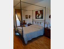 Camera da letto con letto matrimoniale a baldacchino in ferro battuto e copriletto chiaro, affiancato da comodini in legno con lampada. Sulla parete è appeso un grande quadro colorato e a sinistra c’è una portafinestra con tende che dà su un balcone con ringhiera.