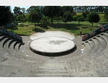 Anfiteatro all’aperto in cemento con gradinate semicircolari e un palco circolare rialzato al centro, con muri laterali decorati da graffiti. Sullo sfondo si vede un’area verde con alberi e prato.