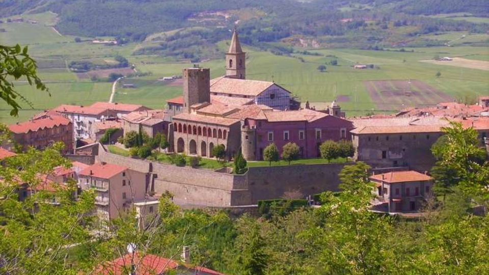 Veduta panoramica di un borgo collinare con complesso religioso in pietra e mattoni, caratterizzato da campanile e torre merlata, racchiuso da mura e circondato da case con tetti in coppi. Sullo sfondo si estendono campi coltivati e colline boscose.