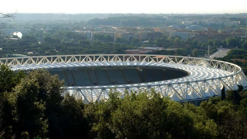 Vista panoramica di un grande stadio ovale con struttura in travi bianche e copertura a membrane, circondato da alberi e aree verdi. Sullo sfondo si vede la città con edifici bassi e strade in lontananza sotto un cielo chiaro.