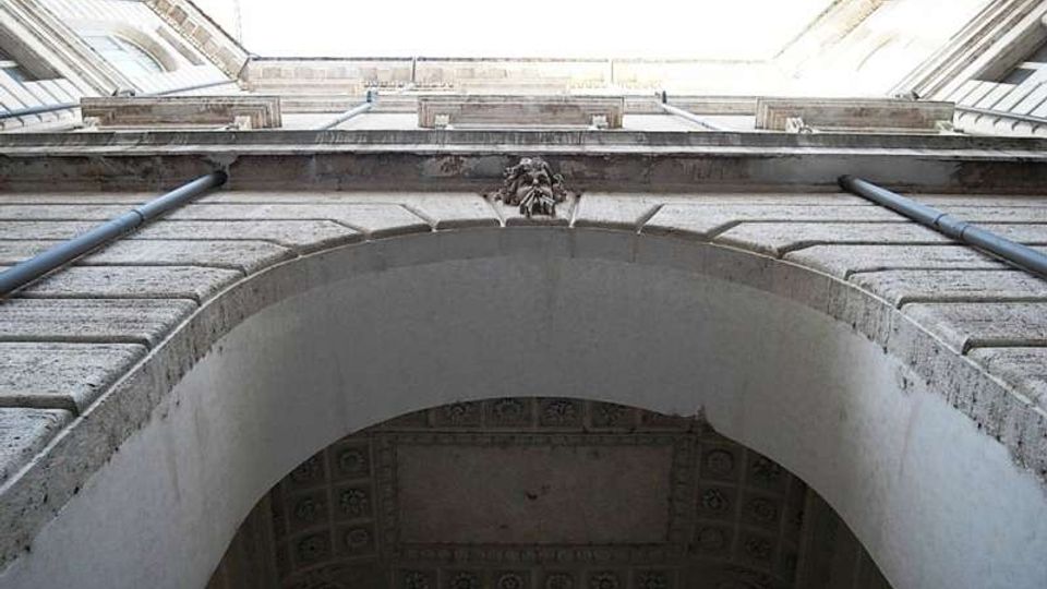 Veduta dal basso di un cortile interno tra edifici in muratura chiara, con grande arco d’ingresso in primo piano e cornici in pietra. Sotto l’arco si intravede un soffitto decorato a cassettoni e lungo le pareti corrono tubazioni verticali.