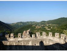 Vista panoramica da una terrazza o camminamento di un castello in pietra, con merlature quadrate in primo piano. Sullo sfondo si vedono colline e vallate verdi con piccoli borghi e campi sotto un cielo sereno.