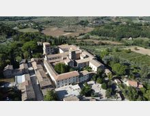 Veduta aerea di un grande complesso monastico in pietra con tetti in coppi, disposto attorno a cortili interni e una torre centrale. L’edificio è circondato da alberi, giardini e campi coltivati in una campagna collinare.