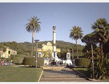 Piazza con ampio prato e scalinata centrale che conduce a un alto monumento su piedistallo, circondato da palme e siepi potate. Sullo sfondo si vedono edifici chiari e una collina boscosa sotto un cielo sereno.