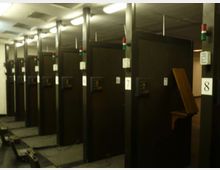 Sala tecnica interna con file di armadiature nere numerate, disposte in corridoi paralleli sotto illuminazione fluorescente a soffitto. Si vedono pannelli di controllo sui lati e una passerella/canalina a pavimento lungo il corridoio.