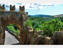 Antica porta in pietra con arco e merli, affacciata su una stretta strada asfaltata con ringhiera. Intorno si vedono rovine di mura e una valle verde con colline sullo sfondo sotto un cielo parzialmente nuvoloso.