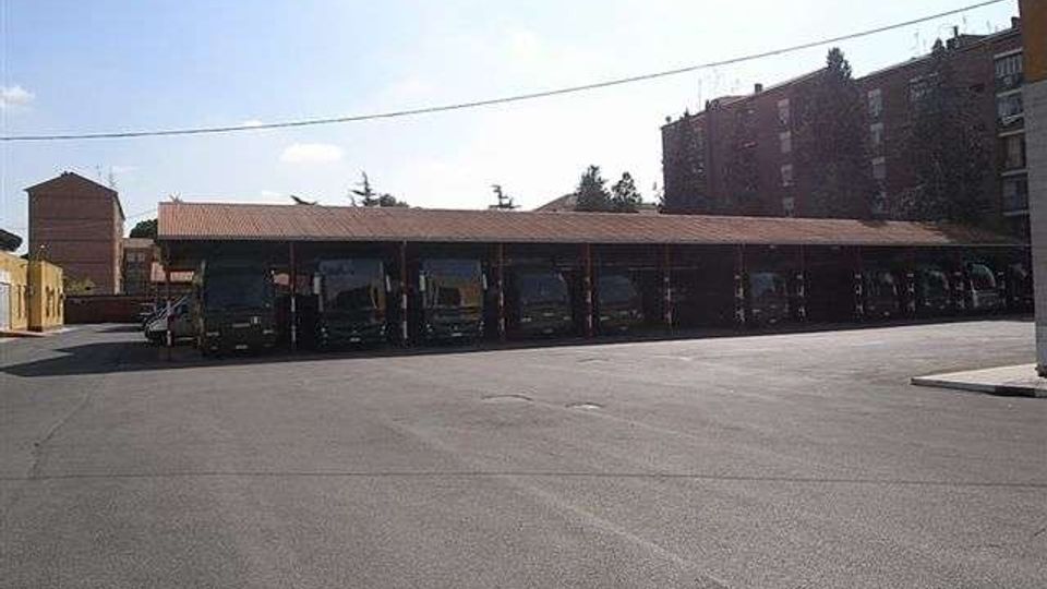 Ampio piazzale asfaltato con una lunga tettoia coperta che ospita una fila di autobus parcheggiati. Sullo sfondo si vedono edifici residenziali in mattoni e alcuni alberi, con cavi elettrici sospesi sopra l’area.