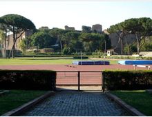 Vista di una pista di atletica all’aperto con pedana per salto in alto e prato interno, delimitata da siepi e un cancello in primo piano. Sullo sfondo si vedono alti pini e mura in pietra di un’area storica.