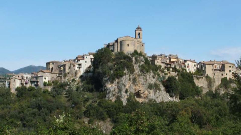 Veduta panoramica di un borgo storico arroccato su uno sperone roccioso, con una chiesa in pietra e campanile in cima e case addossate lungo i fianchi. In primo piano si estende una vegetazione verde, con colline lontane sotto un cielo sereno.