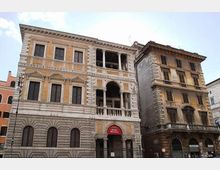 Facciata di un palazzo storico in pietra e intonaco con finestre regolari e loggia centrale ad archi e colonne, con ingresso indicato da un’insegna rossa “Museo Barracco”. Accanto si vede un edificio più stretto e usurato, su una strada urbana con lampione e cielo parzialmente nuvoloso.