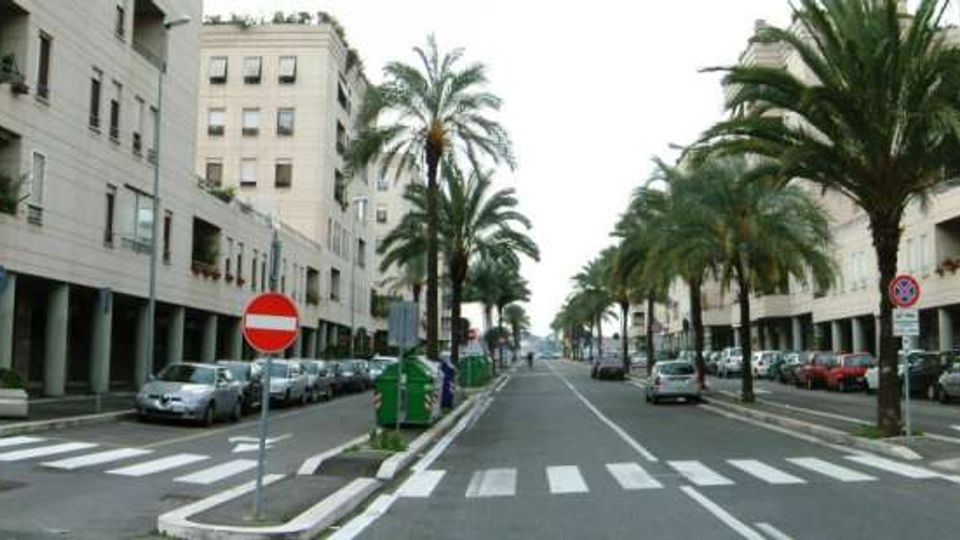 Viale urbano a doppia carreggiata con palme lungo il viale centrale e ai lati, fiancheggiato da edifici moderni con portici al piano terra. Sono visibili strisce pedonali, segnaletica stradale e auto parcheggiate lungo la strada.