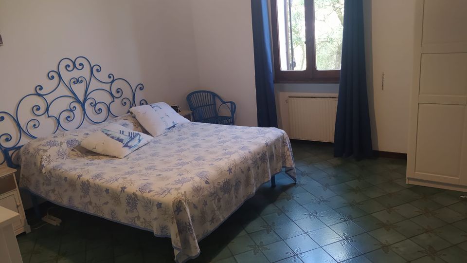 Camera da letto con letto matrimoniale e testiera in ferro battuto blu, copriletto chiaro a motivi marini e pavimento in piastrelle verdi. Una grande finestra ad arco con tende scure illumina la stanza; vicino ci sono una sedia e un armadio bianco.