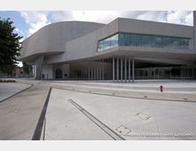 Edificio moderno in cemento grigio con grandi volumi sporgenti e facciata vetrata continua al primo piano, sostenuta da una fila di pilastri sottili. Davanti si apre un’ampia piazza pavimentata in cemento con canalette di drenaggio e pochi elementi urbani, sotto un cielo parzialmente nuvoloso.