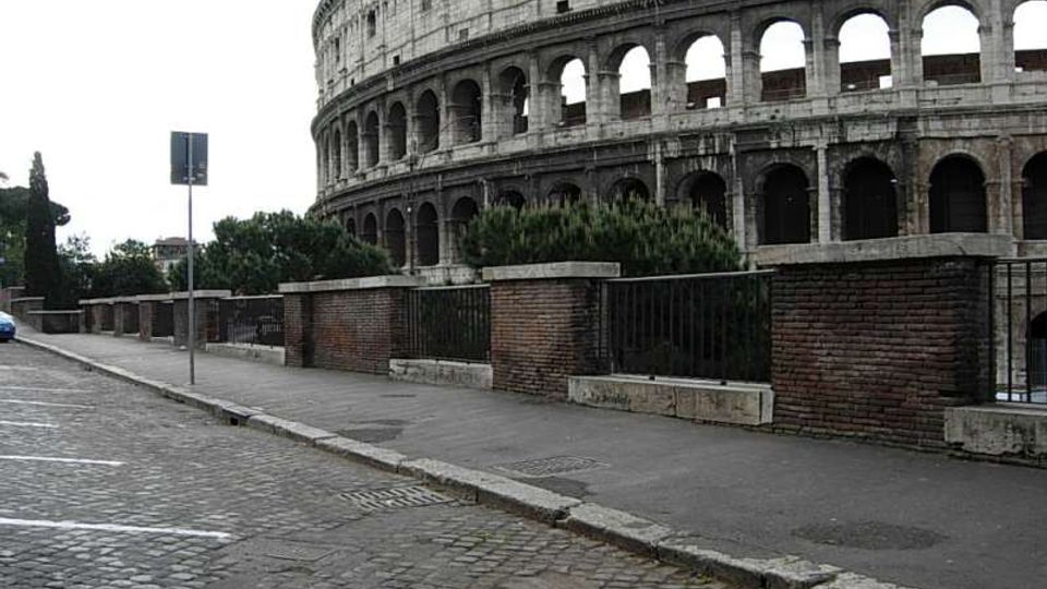 Veduta esterna del Colosseo con le sue arcate in pietra, ripreso da una strada in sampietrini e un marciapiede affiancato da muretti in mattoni e ringhiere metalliche. Sullo sfondo si vedono alberi e un cartello stradale.