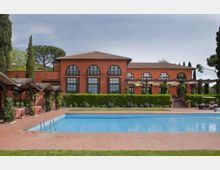 Edificio in mattoni rossi a due piani con tetto in coppi e grandi finestre ad arco, affacciato su un’ampia piscina rettangolare con bordo in cotto. Nell’area esterna ci sono pergolati, siepi curate e alberi alti sullo sfondo.