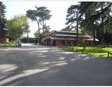 Ampio viale asfaltato in un’area verde con prati, staccionate basse e alti alberi; sullo sfondo si vede un edificio basso con facciata rosa e tetto scuro, affacciato sulla strada.