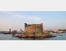 Ruderi di una fortificazione circolare in pietra su un piccolo isolotto roccioso all’ingresso di un porto. Intorno si vedono il mare, moli e strutture portuali con barche ormeggiate sullo sfondo.