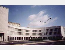 Edificio moderno a più piani con facciata curva e lunga serie di finestre verticali ripetute, affacciato su una strada ampia. In primo piano si vedono un’aiuola centrale e alti lampioni, con cielo azzurro sullo sfondo.