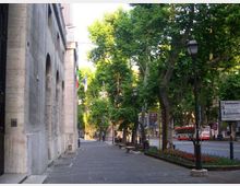 Marciapiede ampio lungo un edificio in pietra con grandi arcate, affacciato su un viale alberato con lampioni. Ai lati si vedono aiuole, panchine e la strada con veicoli sullo sfondo.