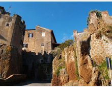 Case in pietra e intonaco di un borgo storico costruite su una parete di roccia tufacea, con resti di mura e vegetazione sulla scarpata. Strada asfaltata in basso e cielo sereno sullo sfondo.