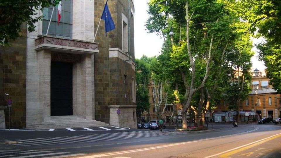 Facciata di un edificio pubblico in pietra con grande portale d’ingresso e bandiere su pennoni, affacciato su una strada con strisce pedonali. Accanto si vede un incrocio alberato con alti alberi e palazzi sullo sfondo.