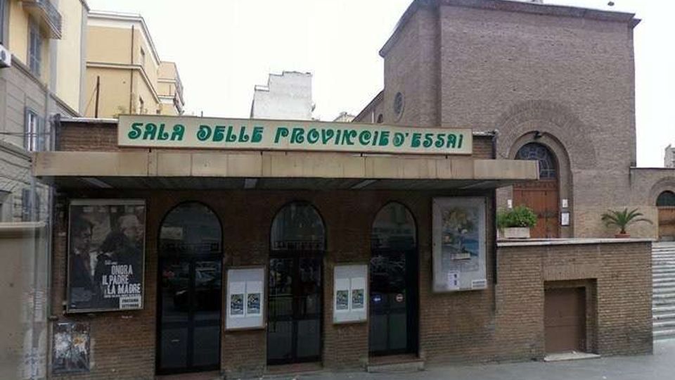Facciata di un edificio in mattoni con ingresso coperto da una pensilina e l’insegna “Sala delle Province d’Essai”. Sotto la tettoia si vedono tre porte ad arco in vetro e bacheche con locandine, con un volume laterale più alto in mattoni e una piccola scala sul lato destro.