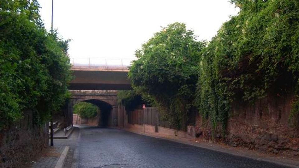 Strada in pavé che passa sotto un ponte e un viadotto, con un sottopasso ad arco in muratura sullo sfondo. Ai lati ci sono muri in mattoni e scarpate ricoperte di vegetazione, con marciapiede e un lampione.