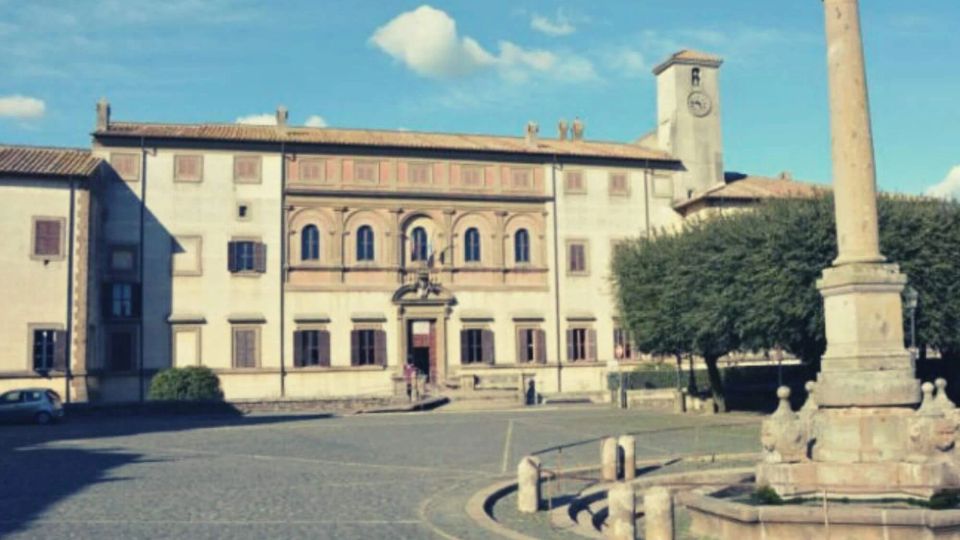 Piazza lastricata con un edificio storico a due piani sullo sfondo, con facciata simmetrica, portale centrale e loggiato ad archi al piano superiore. In primo piano si vede una colonna monumentale su basamento, con alberi a lato e una torre con orologio dietro l’edificio.