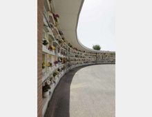 Viale curvo di un cimitero con file di loculi bianchi lungo il lato sinistro, decorati con fiori e targhe, sotto una copertura continua. Pavimentazione in cemento e cielo grigio sullo sfondo.