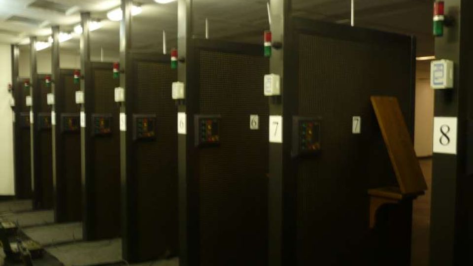 Sala tecnica interna con file di armadiature nere numerate, disposte in corridoi paralleli sotto illuminazione fluorescente a soffitto. Si vedono pannelli di controllo sui lati e una passerella/canalina a pavimento lungo il corridoio.