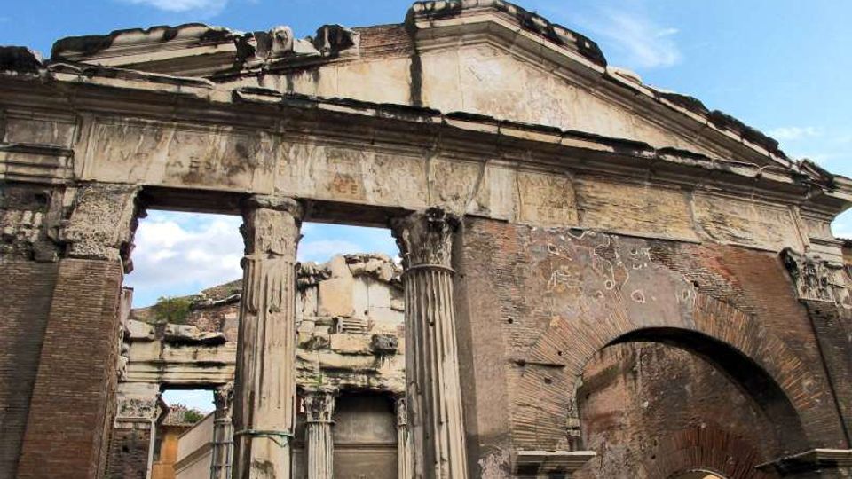 Rovine di una porta monumentale in muratura e pietra, con frontone triangolare e colonne parzialmente danneggiate, affiancata da un grande arco laterale. La struttura appare erosa e incompleta, sullo sfondo di cielo azzurro.
