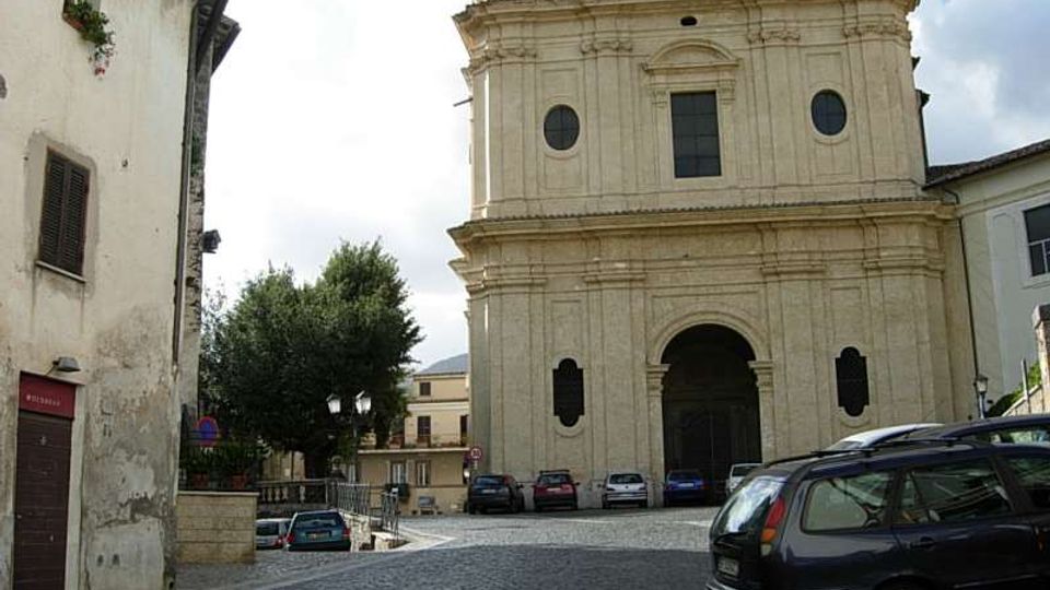 Facciata di una chiesa storica in pietra chiara su una piazza lastricata, con grande portale ad arco centrale, finestre rotonde e frontone classico. Ai lati si vedono edifici residenziali, alcune auto parcheggiate e alberi vicino al margine della piazza.