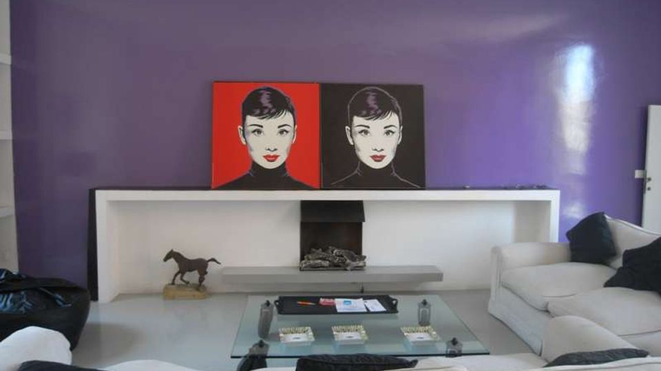 Soggiorno moderno con parete viola e lunga mensola bianca che incornicia un camino centrale. Davanti ci sono un tavolino in vetro e divani chiari, con due quadri pop-art appoggiati sulla mensola e una piccola scultura di cavallo.