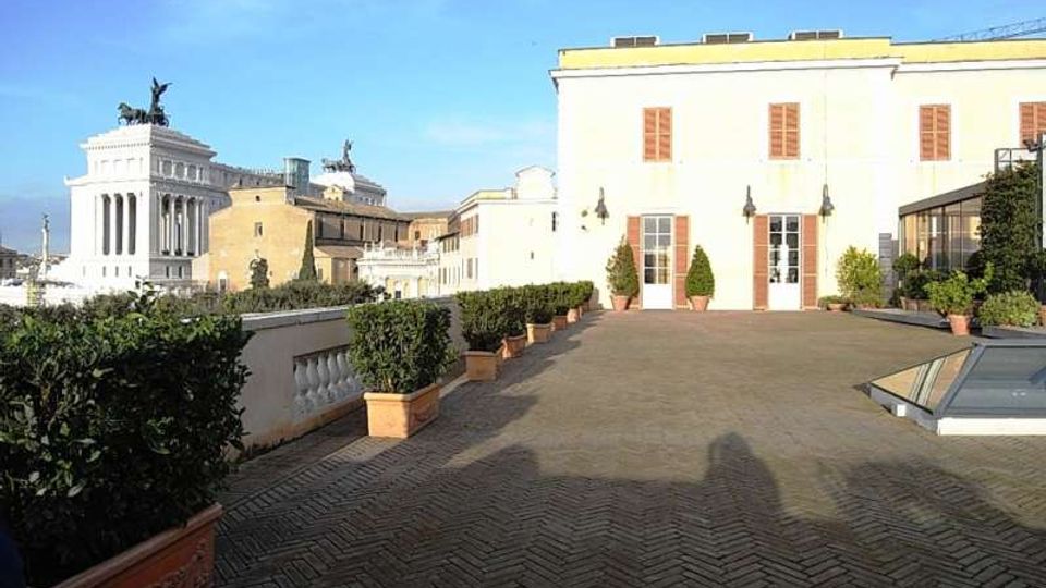 Terrazza panoramica pavimentata in mattoni a spina di pesce con vasi di siepi lungo la balaustra, affacciata su edifici storici. Sullo sfondo si vede un palazzo bianco con persiane in legno e, a sinistra, un grande monumento neoclassico con statue sulla sommità.