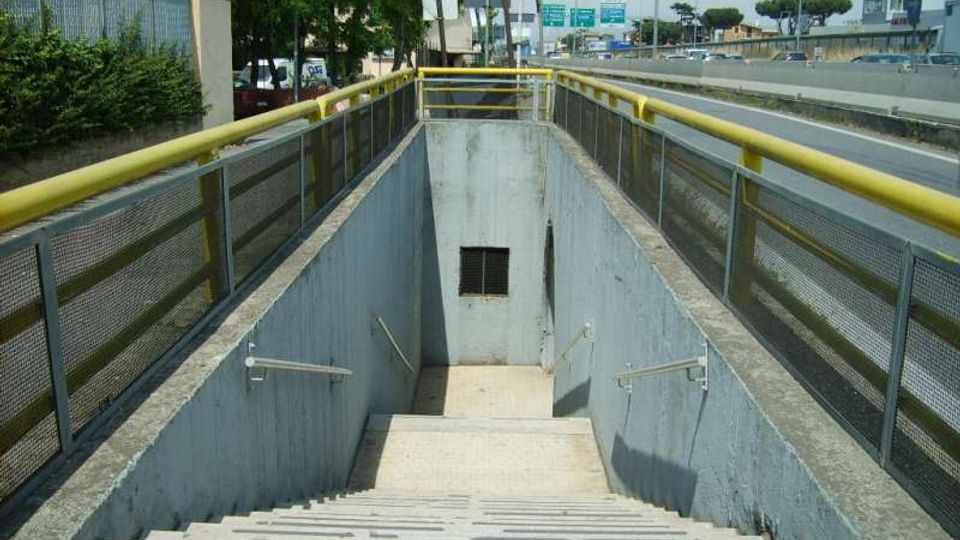 Scala in cemento che scende a un sottopassaggio pedonale, con corrimano laterali e parapetti in rete metallica con fascia gialla. Ai lati si vede una strada a più corsie con guardrail e segnaletica, con alberi e edifici sullo sfondo.