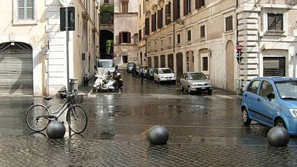 Strada urbana stretta con pavimentazione in pietra bagnata, fiancheggiata da edifici storici in muratura con finestre e archi al piano terra. In primo piano si vedono dissuasori sferici, una bicicletta e veicoli parcheggiati lungo i lati della via.