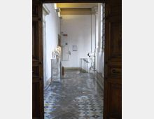 Interno di un edificio storico visto attraverso un grande portone in legno, con pavimento a mosaico geometrico e pareti bianche. Lungo il corridoio si notano una scultura su piedistallo, colonne e finestre alte, e una ringhiera che delimita una scala.