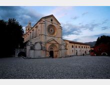 Facciata di una grande chiesa in pietra con rosone centrale e portale ad arco, affacciata su un’ampia piazza lastricata. A lato si estende un lungo edificio adiacente, con alberi e colline sullo sfondo sotto un cielo nuvoloso.