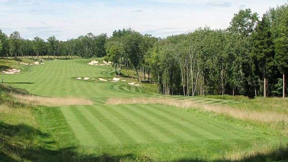 Campo da golf con fairway ampio e ondulato, delimitato da erba alta e circondato da un fitto bosco di alberi. Lungo il percorso si vedono diverse buche di sabbia bianche e leggere pendenze del terreno sotto un cielo parzialmente nuvoloso.