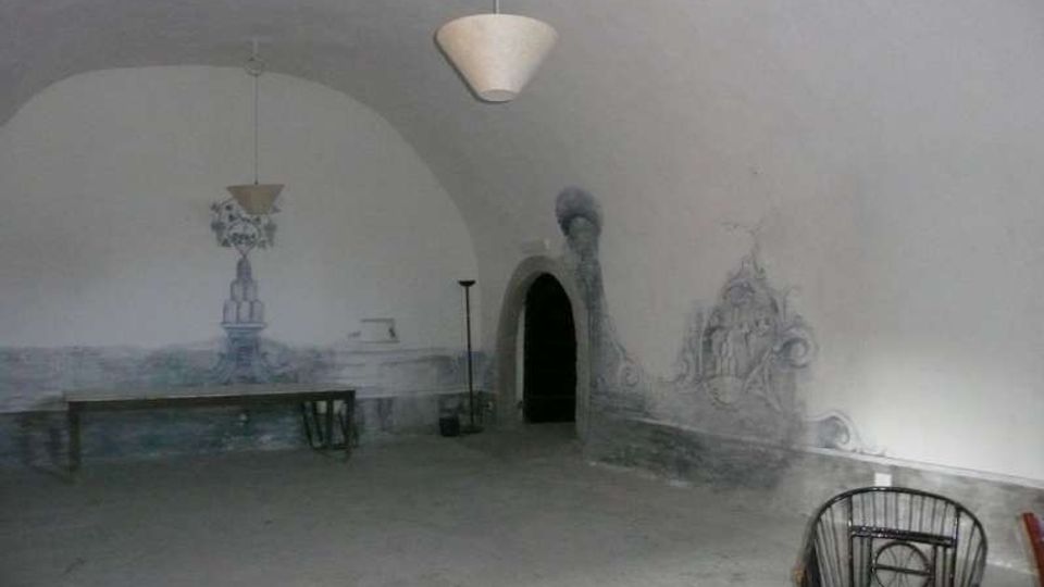 Ampio ambiente interno con soffitto a volta e pareti bianche decorate da affreschi sbiaditi nella fascia inferiore. Al centro si vede un’apertura ad arco scura e l’arredo è minimale, con una lunga panca e alcune sedie, e lampade a sospensione dal soffitto.