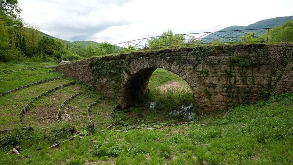 Ponte in pietra con un’unica arcata sopra un piccolo corso d’acqua, con parapetto metallico sulla sommità. Intorno si vede una valle erbosa con binari ferroviari dismessi curvi, alberi e colline sullo sfondo sotto un cielo nuvoloso.