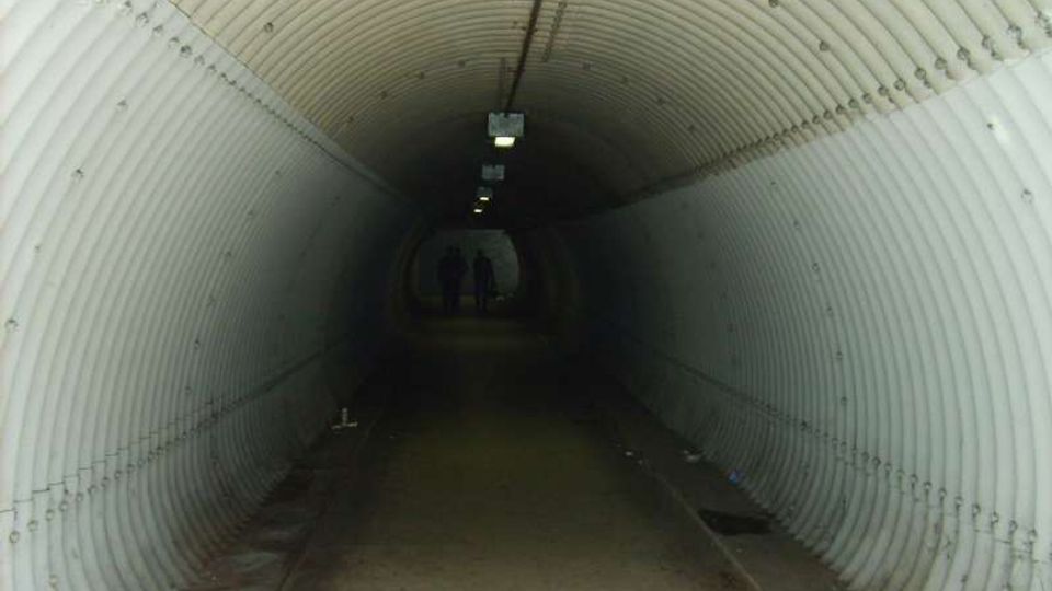 Tunnel pedonale sotterraneo con pareti e soffitto semicircolari in pannelli metallici ondulati, illuminato da una fila di lampade a soffitto. Il pavimento in cemento corre dritto verso un’uscita luminosa in fondo al passaggio.