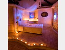 Camera da letto in stile romantico con grande letto matrimoniale, baldacchino in tessuto bianco e illuminazione soffusa a candele. Pavimento piastrellato con gradino curvo, porte in legno e parete decorativa in vetrocemento con luce integrata.