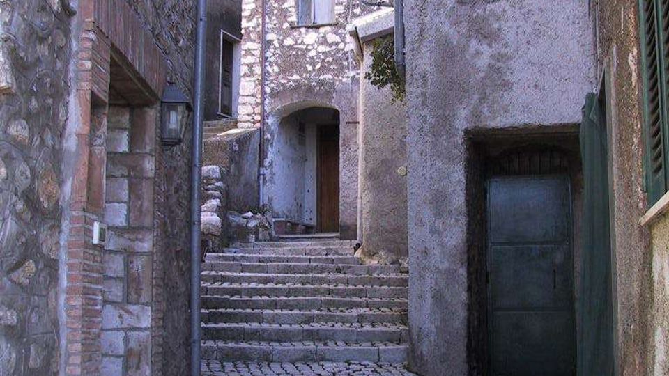 Vicolo stretto in un borgo antico con pavimentazione in ciottoli e gradini in pietra che salgono tra case in muratura e intonaco. In fondo si vede un arco con portone e piccole finestre, con pareti in pietra a vista e persiane.