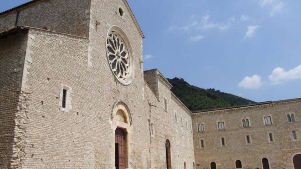 Facciata di una chiesa in pietra chiara con grande rosone circolare e portale ad arco raggiunto da alcuni gradini, affacciata su un ampio cortile. Sul lato destro si estende un edificio in pietra con finestre regolari e archi al piano terra, con colline verdi e cielo azzurro sullo sfondo.