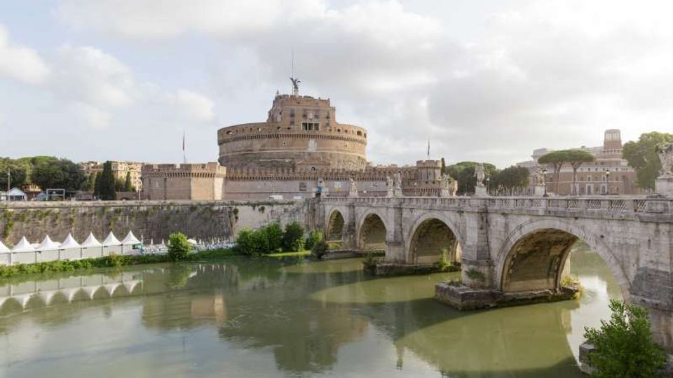 Veduta di Castel Sant’Angelo a Roma, grande fortezza cilindrica in pietra, con il Ponte Sant’Angelo ad archi in primo piano che attraversa il fiume Tevere. Sullo sfondo si vedono mura, edifici storici e alberi lungo le rive sotto un cielo parzialmente nuvoloso.