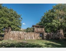 Recinto in legno con pali verticali e un portale centrale sormontato da una piccola torretta coperta, in un’area erbosa. Grandi alberi circondano la struttura e sullo sfondo si vede una collina sotto un cielo sereno.