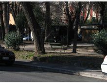 Area verde urbana con alberi alti e cespugli, attraversata da un vialetto con panchine. Sullo sfondo si vedono auto parcheggiate e edifici residenziali dietro la vegetazione.