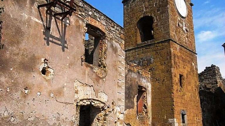 Rovine di un edificio in pietra e intonaco con muri parzialmente crollati e aperture senza infissi, accanto a una torre campanaria quadrata in pietra con grandi arcate e un quadrante circolare. La scena è in una piazzetta lastricata, con detriti e resti di strutture metalliche sotto un cielo azzurro.